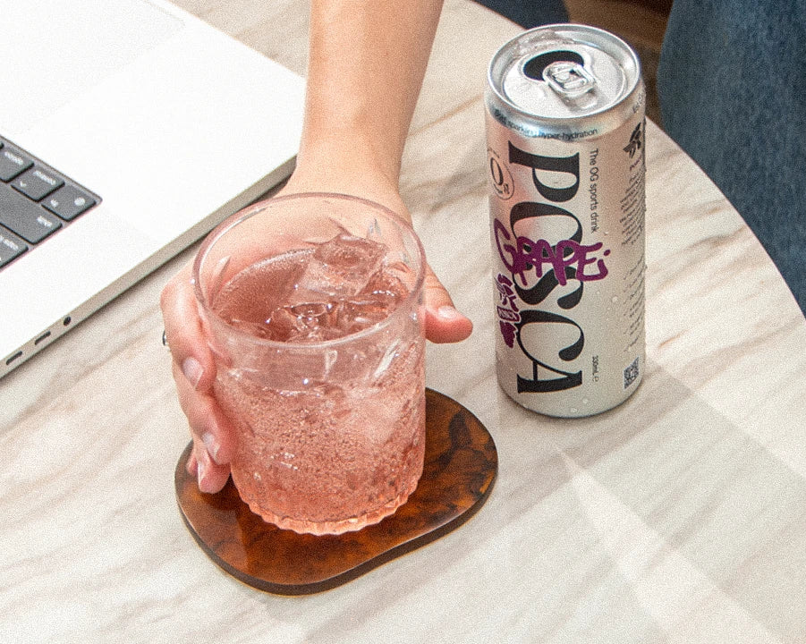 Person holding a Posca Hydrate Grape in a glass with a can on a table next to a laptop.